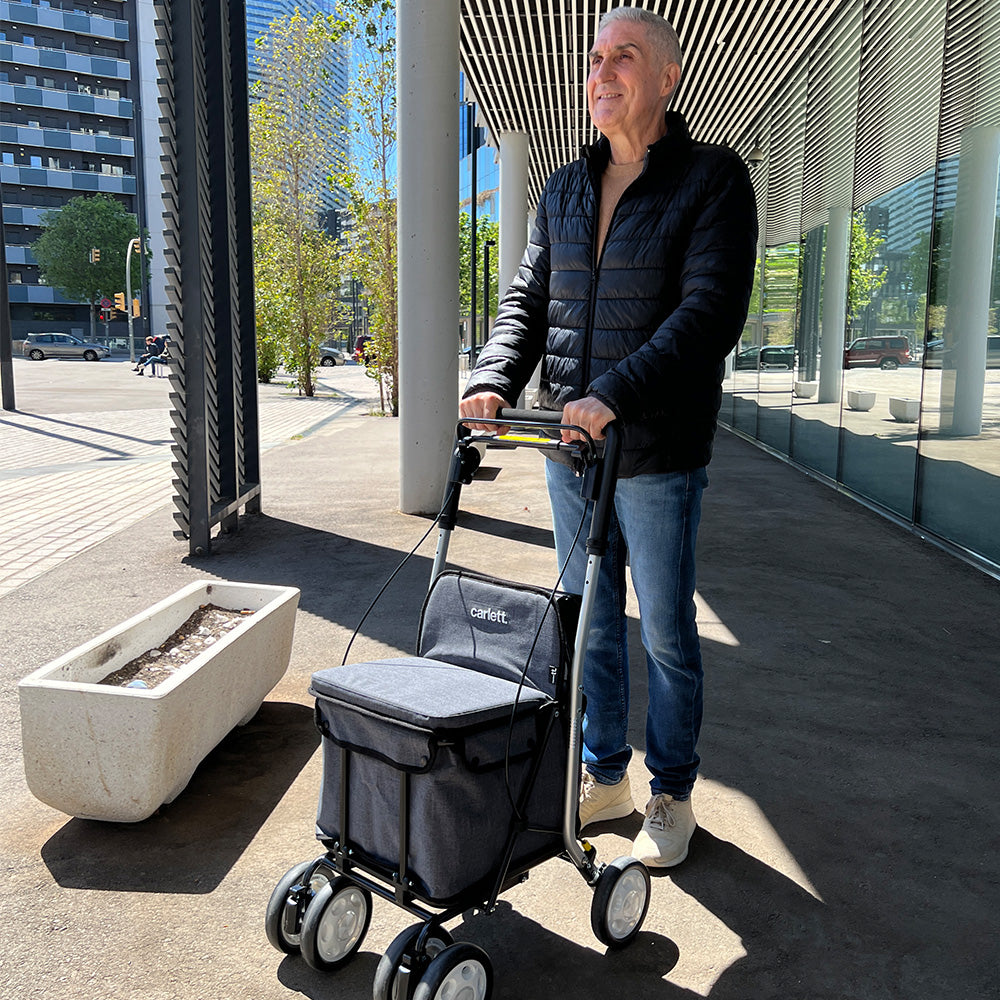 Person steht unter einem Dachvorsprung in einer Stadt und hält den Einkaufsrollator comfort Move in Grau in den Händen.