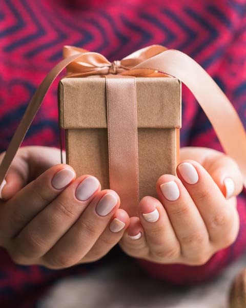 Hände einer Frau, die im Vordergrund ein eingepacktes Geschenk mit Schleife hält.