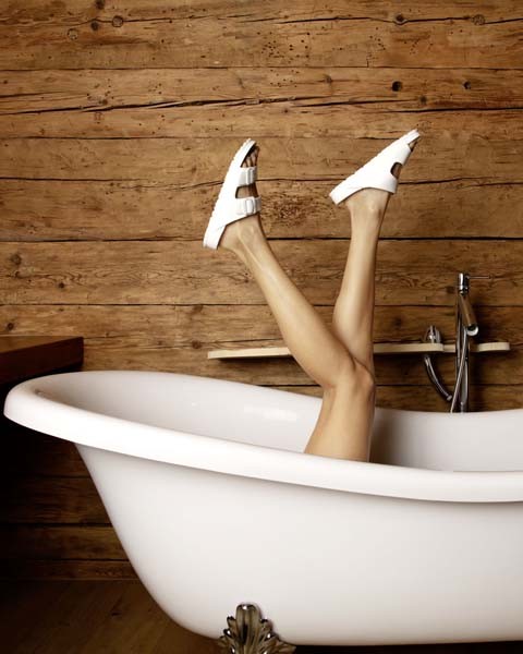 Beine einer Frau mit Birkenstock® Schuhe in einer weißen Badewanne vor Holzhintergrund