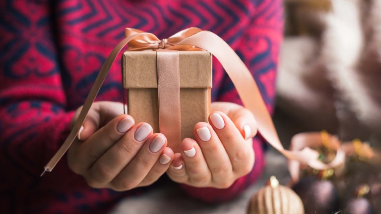 Hände einer Frau, die im Vordergrund ein eingepacktes Geschenk mit Schleife hält.
