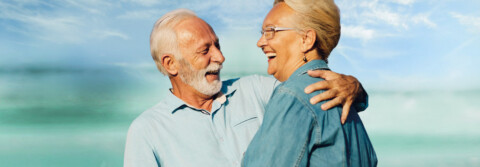 Älteres Paar lächelt sich an; der Mann umarmt die Frau vor einem unscharfen blauen Hintergrund.
