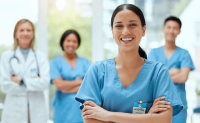 Lächelnde Krankenschwester in blauer Dienstkleidung im Vordergrund; unscharfes medizinisches Team im Hintergrund.