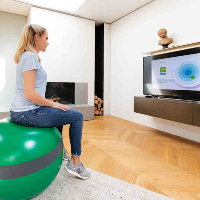 Frau sitzt auf MFT® Balance Sensor Sit Ball vor Fernseher im Wohnzimmer
