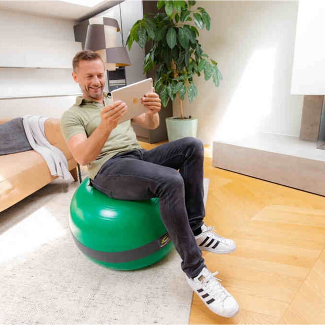 Mann sitzt auf MFT® Balance Sensor Sit Ball und hält ein Tablet für digitales Rückentraining im Sitzen