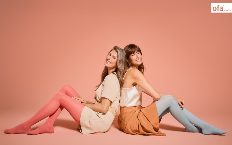 Zwei lächelnde Frauen sitzen Rücken an Rücken vor pfirsichfarbenem Hintergrund und tragen pastellfarbene Stützstrümpfe.