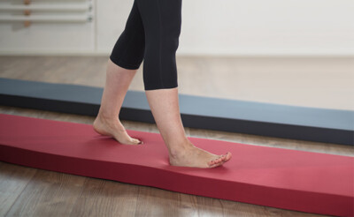 Person geht barfuß über eine rote Balance- oder Stehmatte in einem Innenraum.