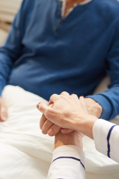 Alter Herr liegt in Pflegebett und hält Hände mit seiner Partnerin.