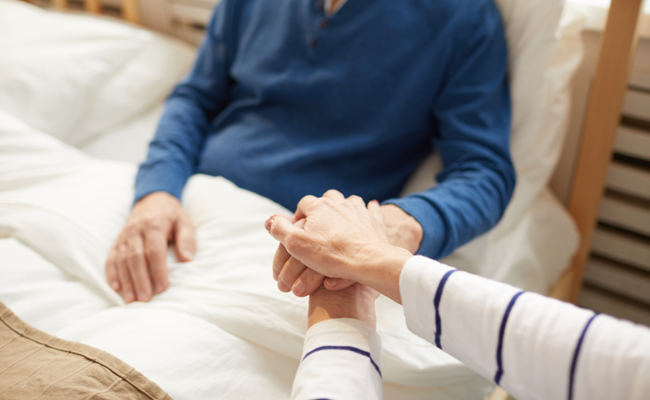 Alter Herr liegt in Pflegebett und hält Hände mit seiner Partnerin.