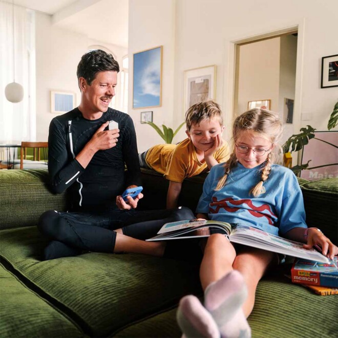Vater sitzt mit seinen Kinder auf couch und lest Buch, während er den Exopulse Suit trägt.