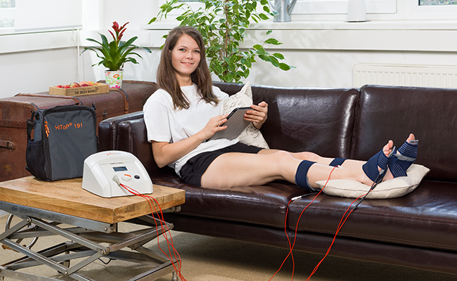 Frau sitzt auf Sofa und hat Elektroden an Ihren Füßen.