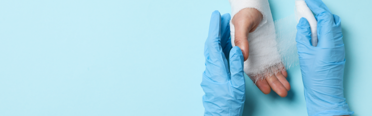 Zwei Hände die Handschuhe tragen versorgen eine andere Hand mit einem Verband auf blauem Hintergrund.