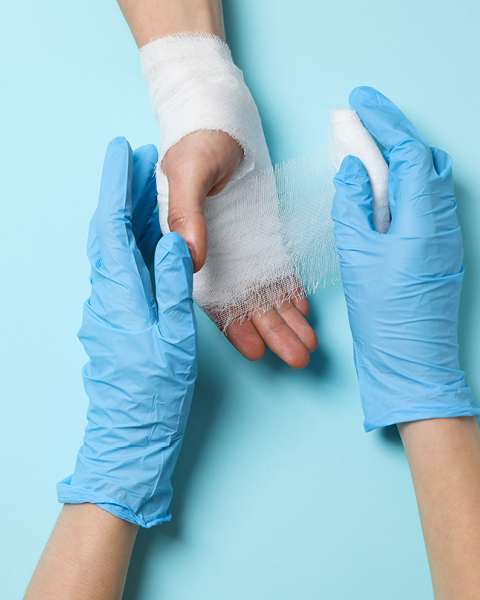 Zwei Hände die Handschuhe tragen versorgen eine andere Hand mit einem Verband auf blauem Hintergrund.