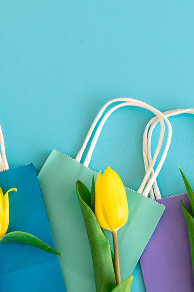 Collage auf hell blauem Hintergrund mit bunten Einkaufstüten und gelben Tulpen.